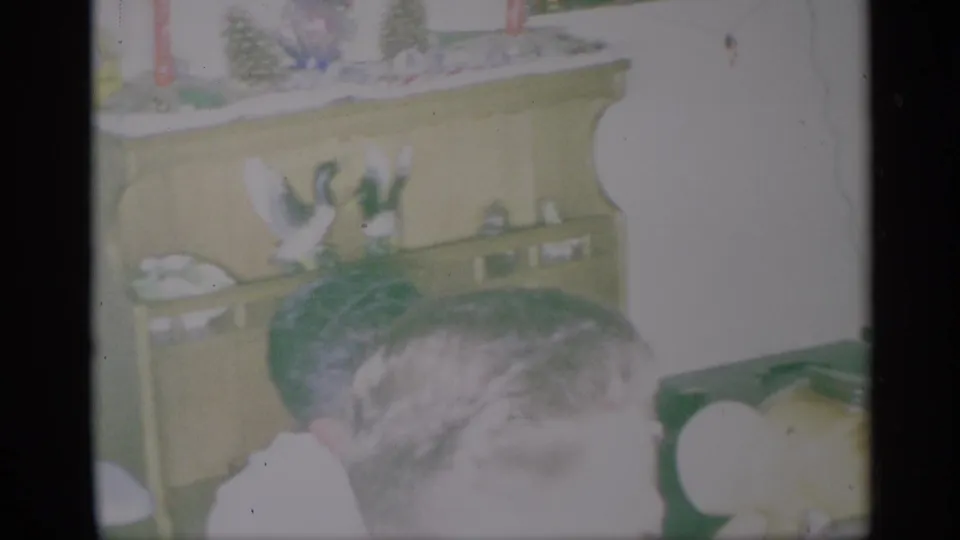 1959 New York home-movie frame showing adults near a hutch with ceramic birds and holiday decorations.