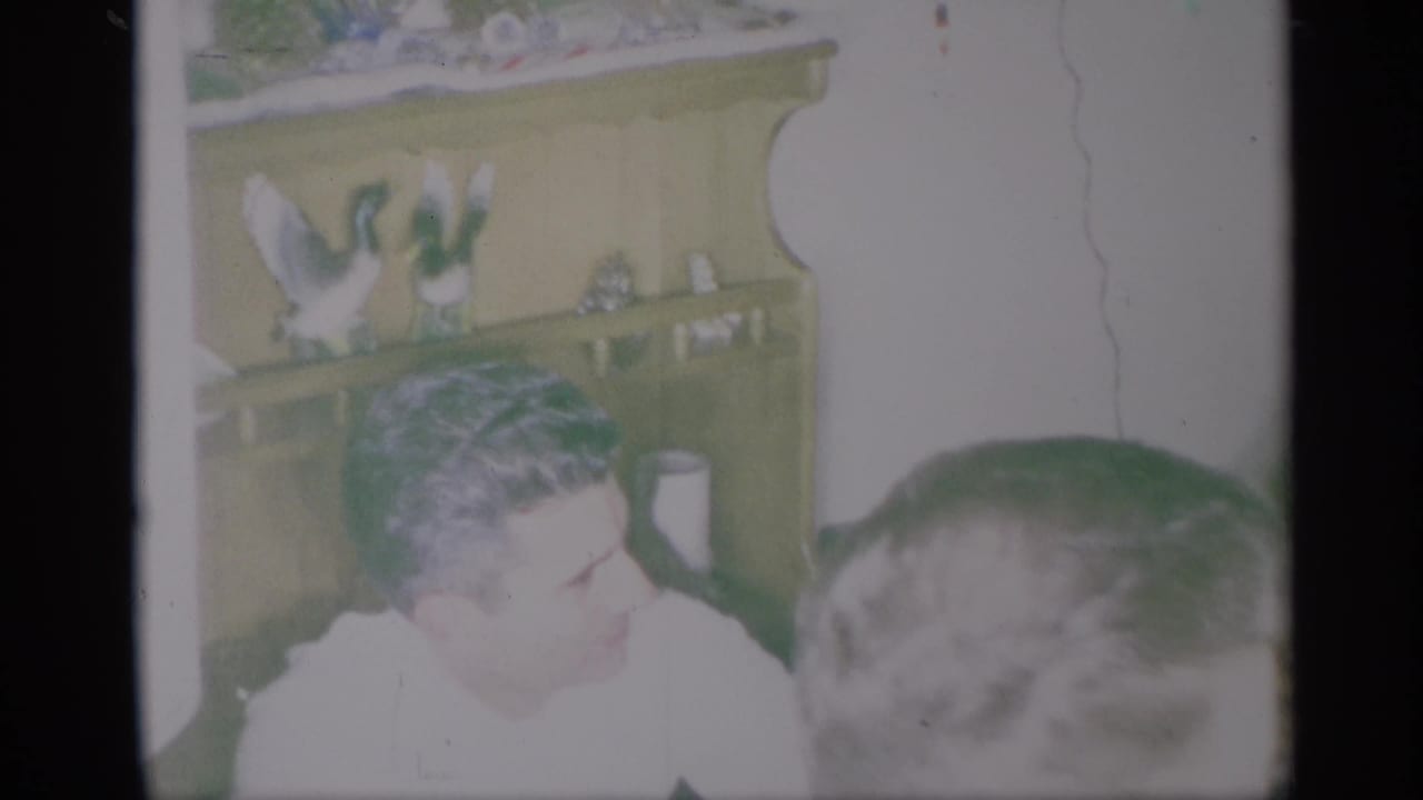 A seated man in a white shirt faces another person beside a decorated hutch.