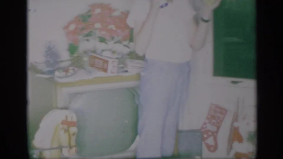 A standing adult is partly framed beside a television, flowers, and holiday items in the 1959 New York home interior.