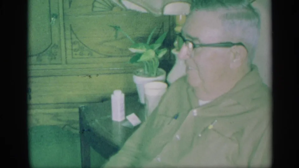 In a 1959 New York home-movie frame, a man in glasses sits near a lamp, plant and table inside a private home.