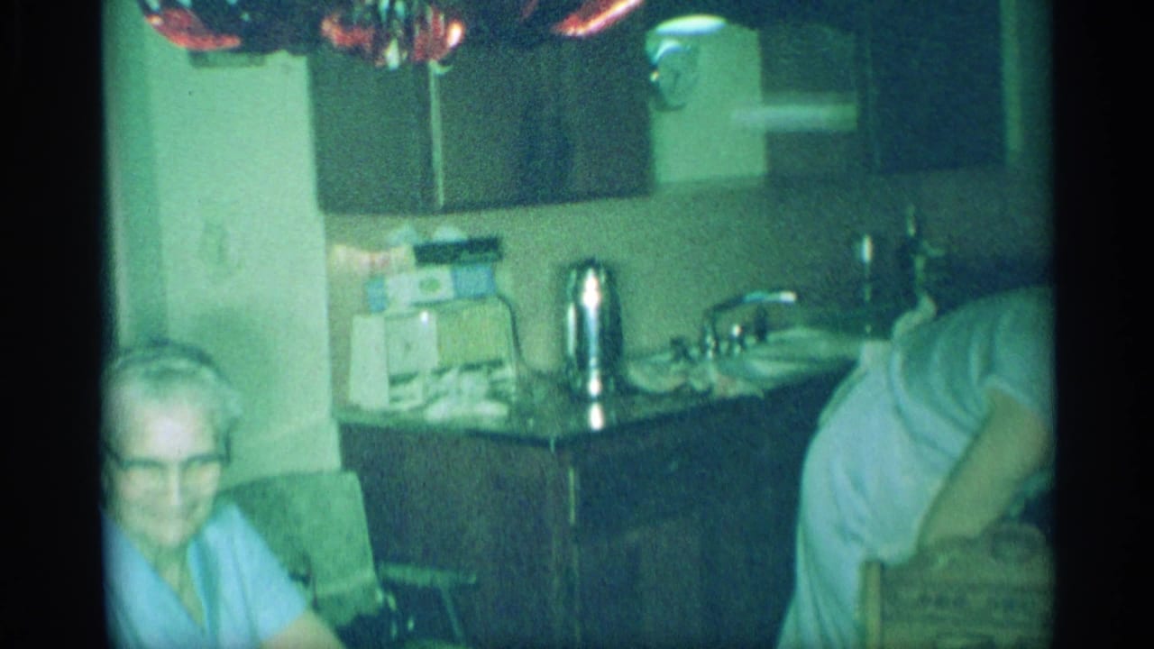 Older man seated at left while a woman bends near a kitchen counter.