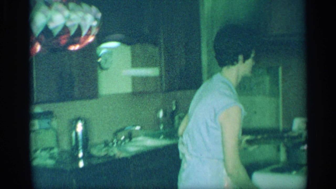 1959 New York home-movie frame with a woman in a light dress standing by a sink and counter under a hanging kitchen light.