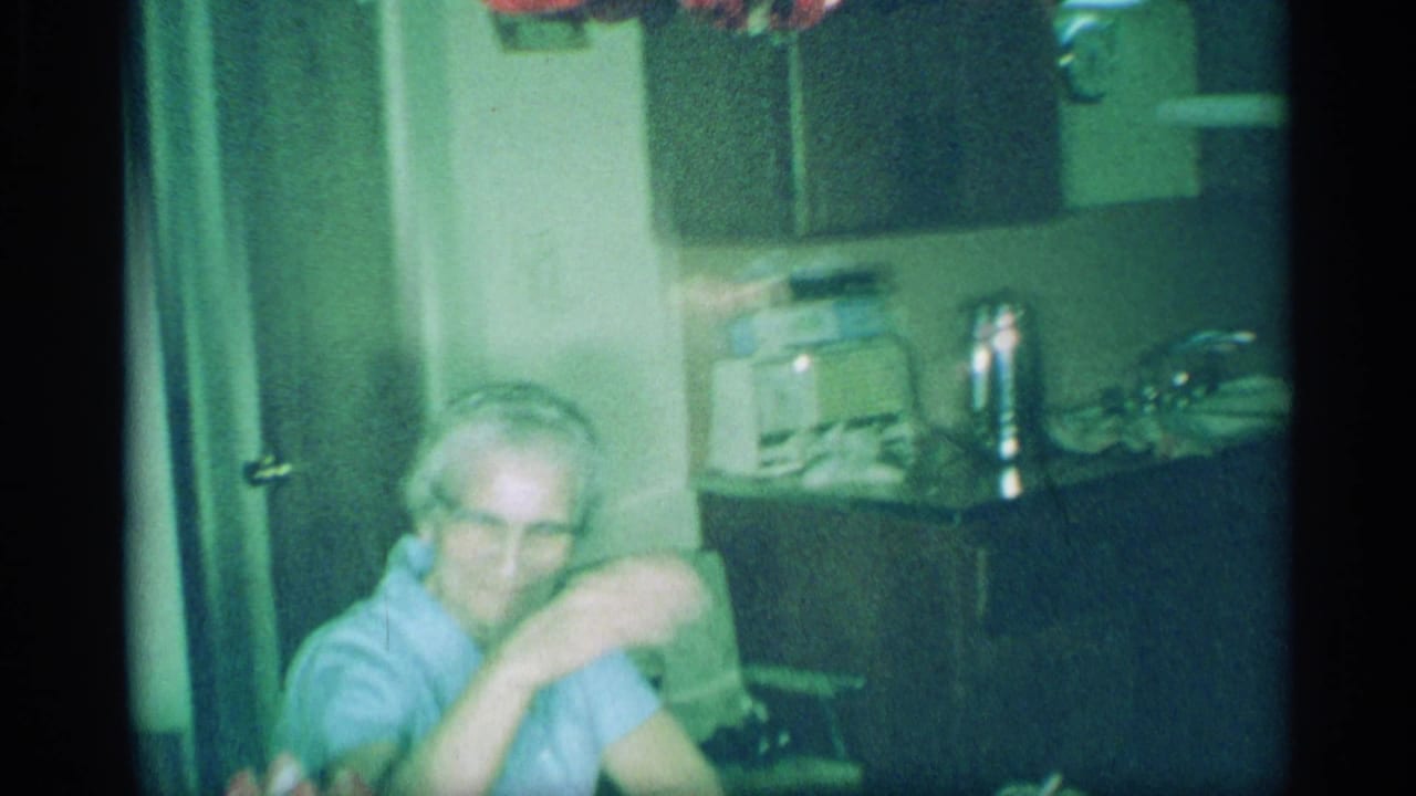 1959 New York home-movie frame showing an older man with glasses seated at a table beside the kitchen counter and appliances.