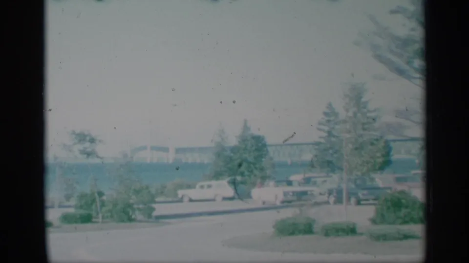 1959 New York home-movie frame showing classic cars parked near a lakeside park road with trees, shrubs, and a bridge beyond the water.
