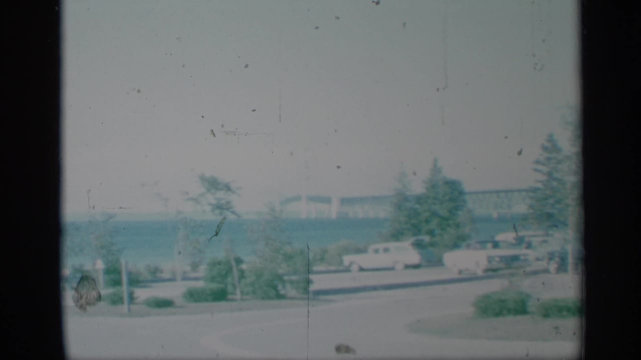 1959 New York lakeside park view with parked mid-century cars along a road and a long bridge visible across the water.