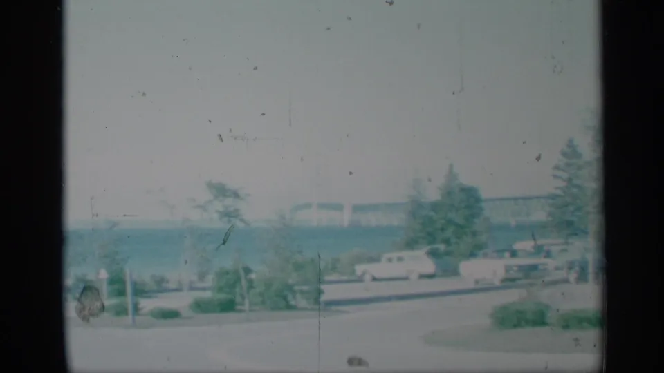 1959 New York lakeside park view with parked mid-century cars along a road and a long bridge visible across the water.