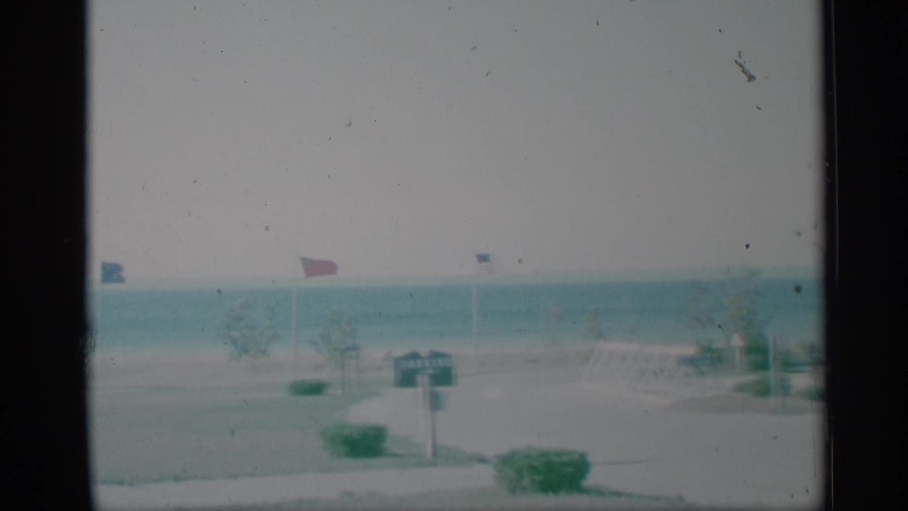 1959 New York home-movie frame of a waterfront park lawn with small flags, shrubs, a signpost, and open water in the background.