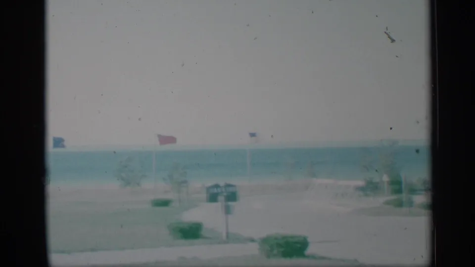 1959 New York home-movie frame of a waterfront park lawn with small flags, shrubs, a signpost, and open water in the background.