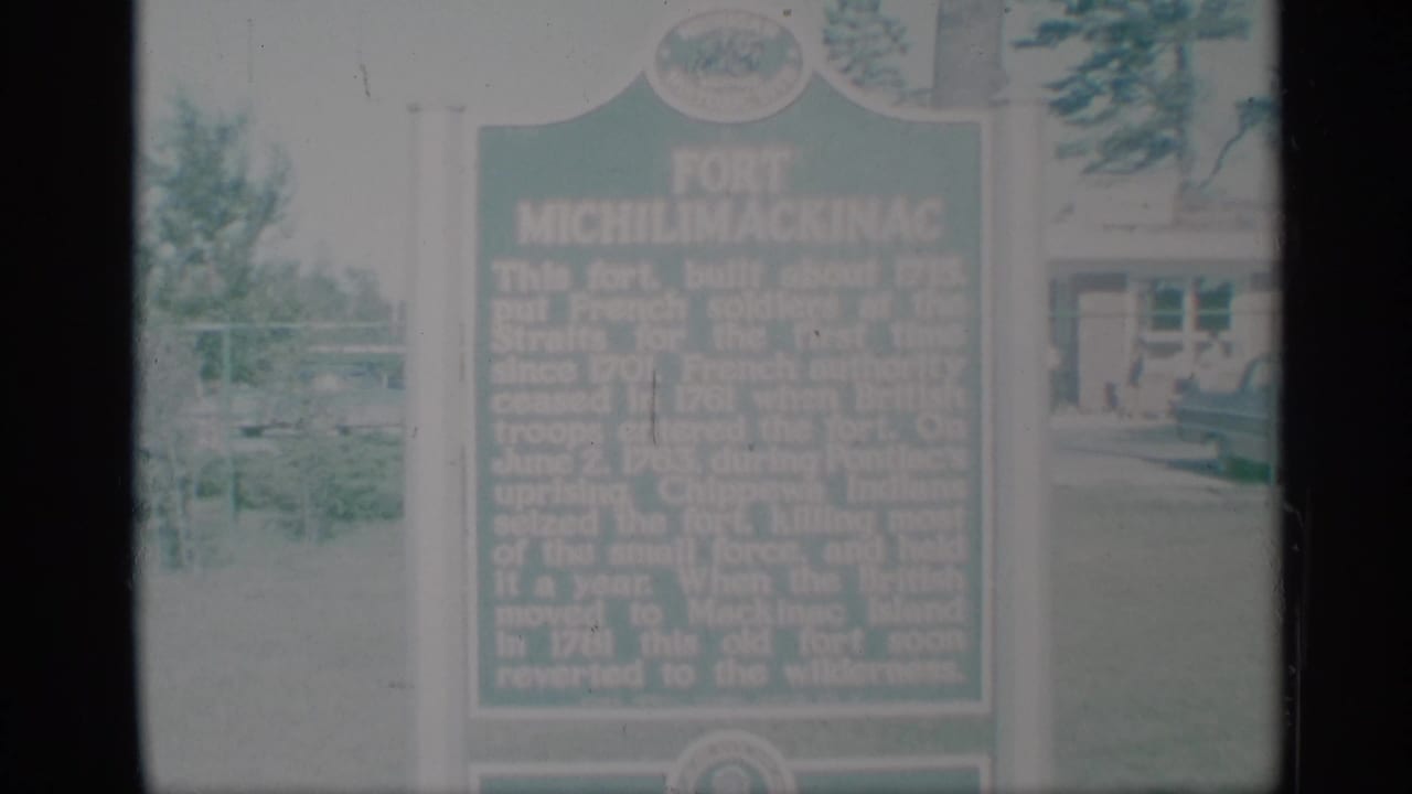 1959 8mm frame showing a green Fort Michilimackinac historical marker in grass with trees and a building behind it.