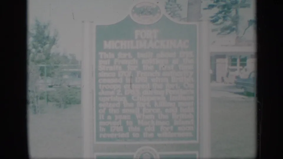 1959 8mm frame showing a green Fort Michilimackinac historical marker in grass with trees and a building behind it.