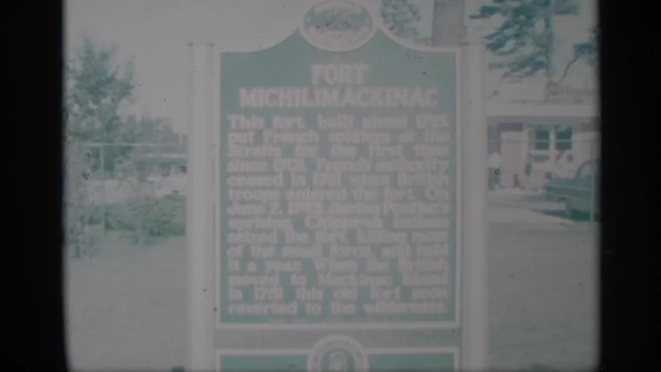 The marker remains centered in a faded 1959 8mm view, with trees, lawn, and small distant visitors near a background building.