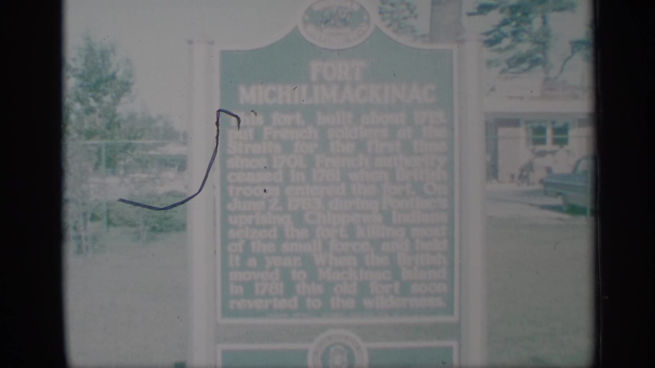 A soft 1959 8mm view of the green marker, grass, and building, with a visible film scratch across the left portion of the frame.