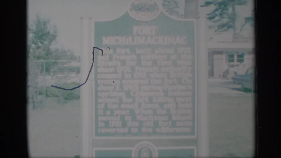 A soft 1959 8mm view of the green marker, grass, and building, with a visible film scratch across the left portion of the frame.