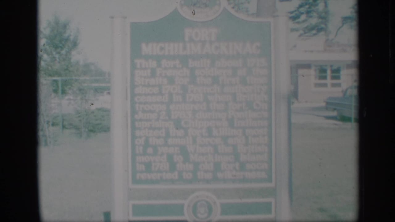 Green Fort Michilimackinac historical marker outdoors with trees and a building behind it.