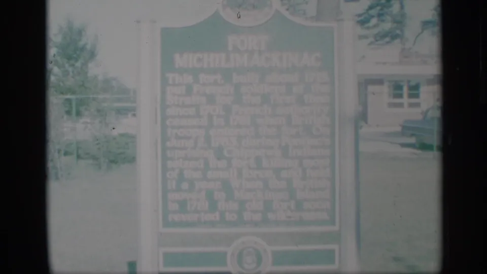 1959 8mm frame of a Fort Michilimackinac historical marker, softly focused and overexposed in an outdoor visitor setting.