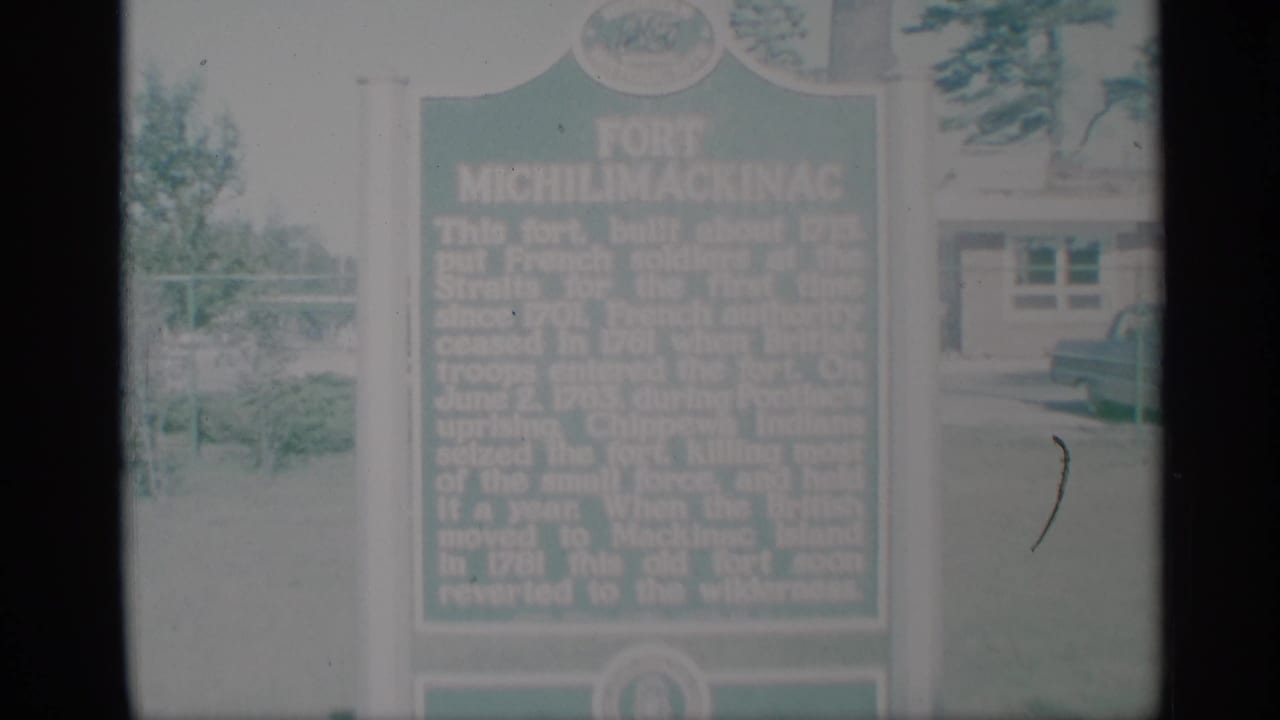 Soft 1959 home-movie view of the Fort Michilimackinac marker, with background building details and pale outdoor exposure.