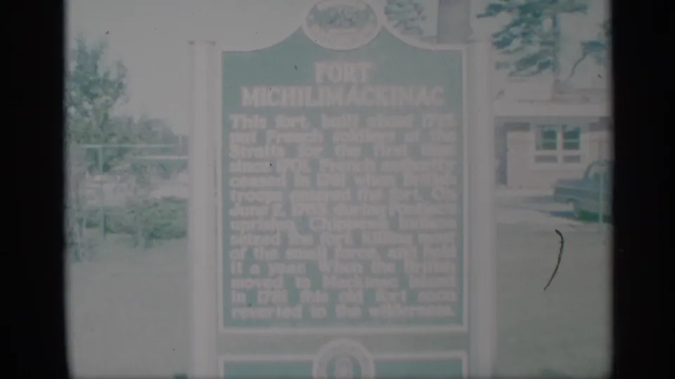 Soft 1959 home-movie view of the Fort Michilimackinac marker, with background building details and pale outdoor exposure.