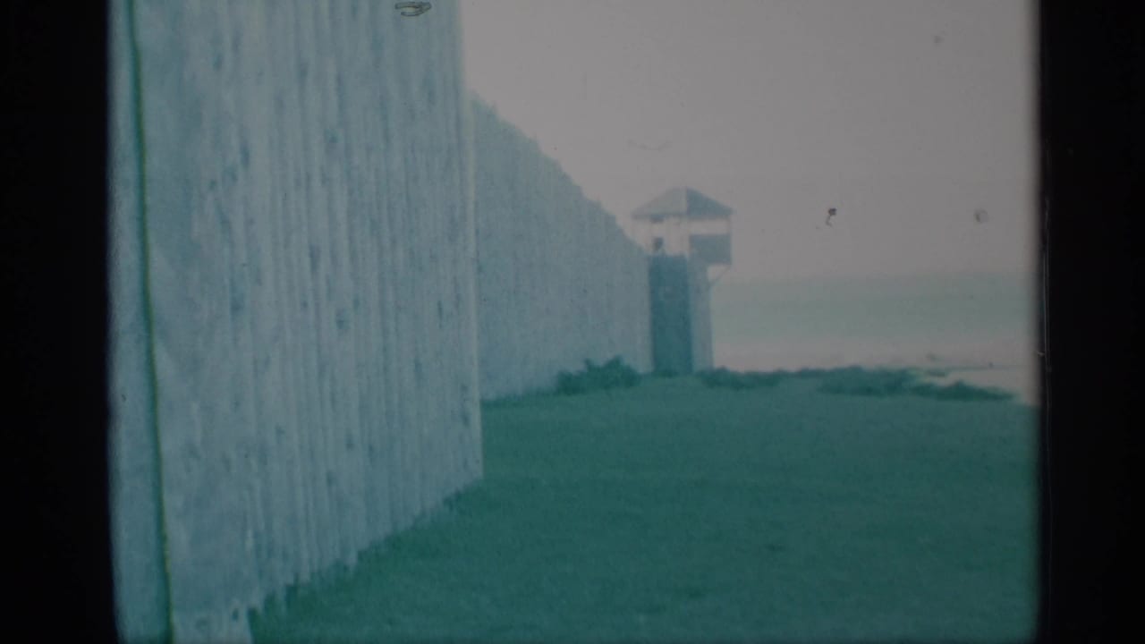 Visitor-shot 8mm view of a wooden palisade wall and distant guard tower at a preserved historic site in 1959.