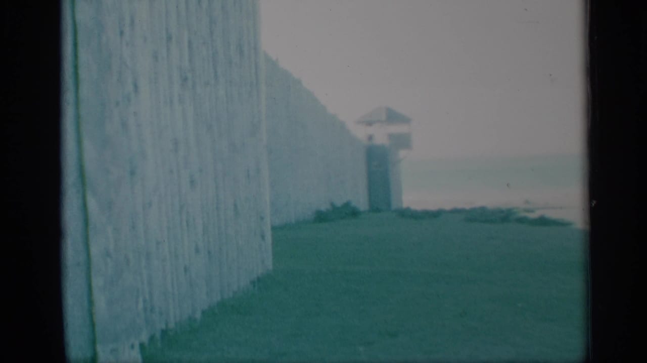 Faded 8mm frame showing a palisade-like wooden wall, grass, and a small guard tower beyond it at the historical site.