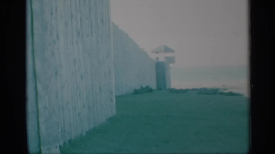 Faded 8mm frame showing a palisade-like wooden wall, grass, and a small guard tower beyond it at the historical site.