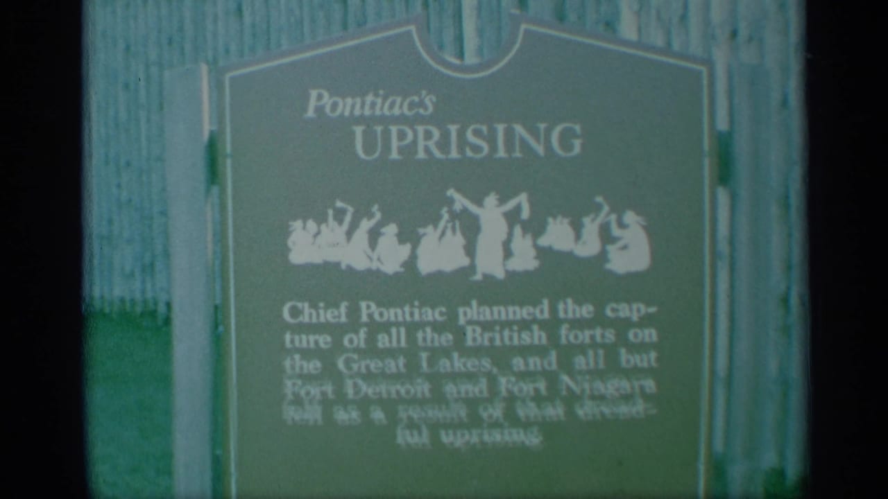 Soft 8mm frame of a green plaque titled Pontiac's Uprising beside a wooden fence.