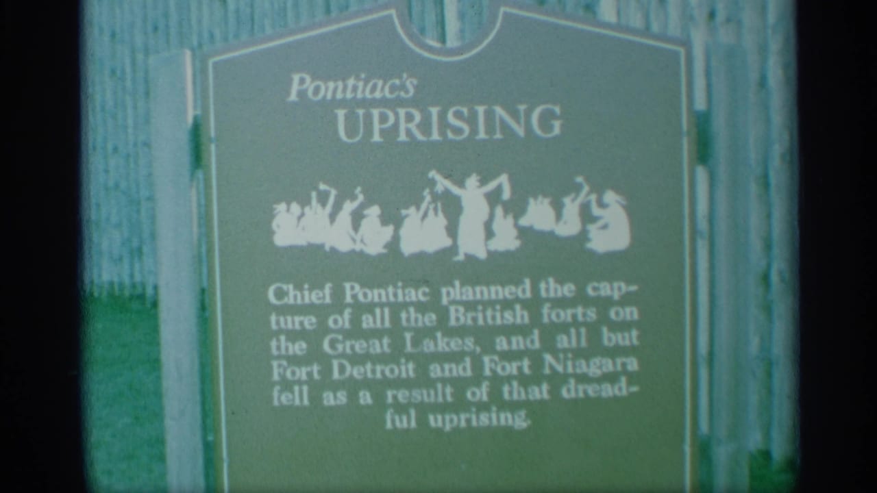 Close visitor-shot 8mm view of the Pontiac's Uprising plaque in New York, with readable title, white silhouette art, and fence boards behind it.