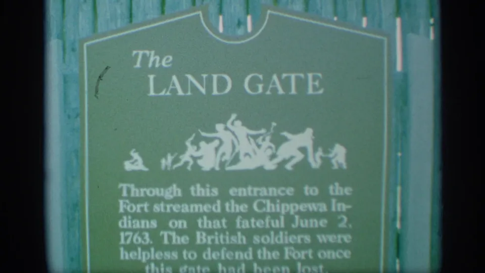 1959 New York home-movie frame shows The Land Gate marker at a fort, with white lettering, silhouette art, and text mentioning Chippewa Indians.