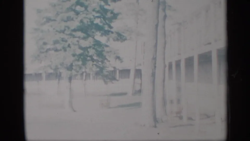 Faded 1959 New York 8mm frame showing a low building, bare trees, open ground, and a covered walkway.