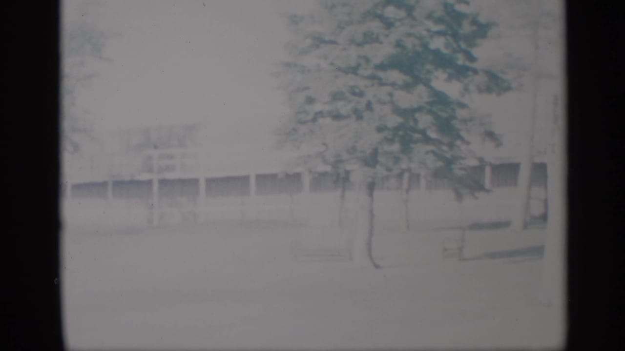 Washed-out 1959 New York home-movie frame with a horizontal building facade, scattered trees, and open foreground.