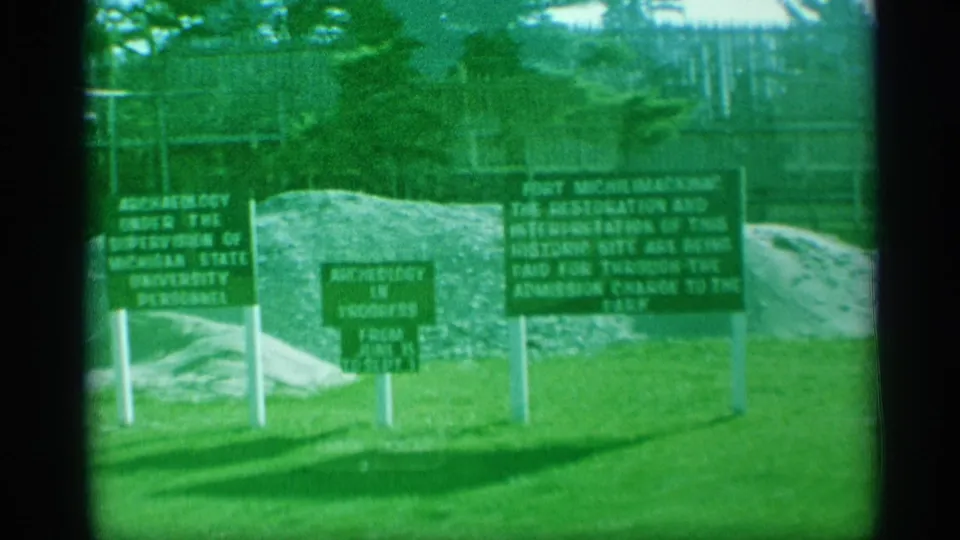 Visitor-shot 8mm frame of Fort Michilimackinac signage describing archaeology, restoration, and admission-supported work.