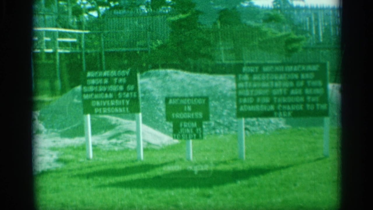 Soft green-tinted 8mm view of signs on historic fort grounds with excavated earth and a stockade fence behind.