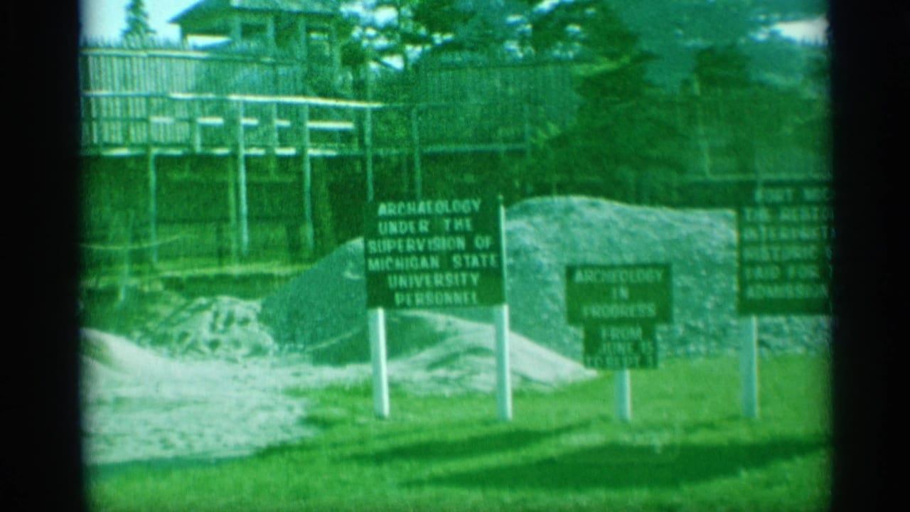 1959 home-movie frame showing archaeology signage near a raised walkway and wooden fence at the historic fort site.
