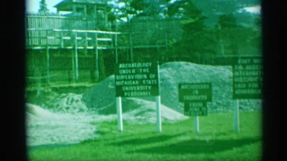1959 home-movie frame showing archaeology signage near a raised walkway and wooden fence at the historic fort site.