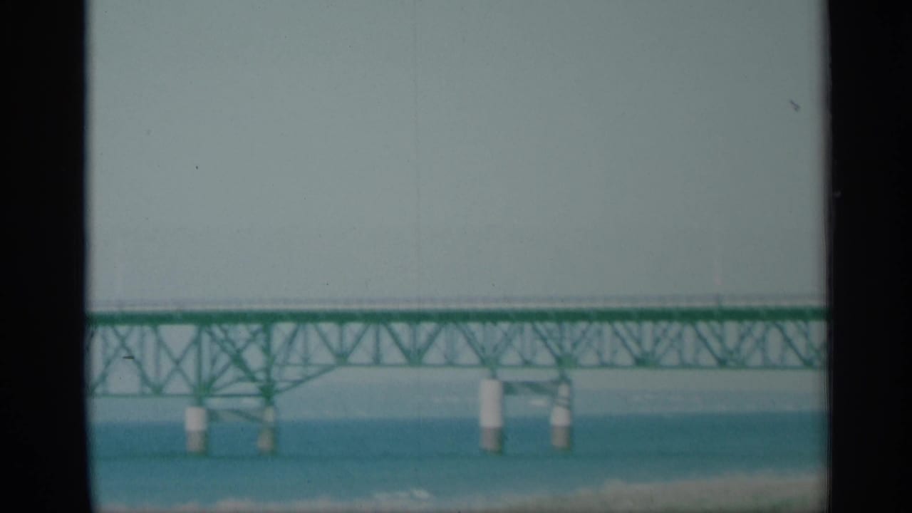 Distant green truss bridge over blue water with dark film-frame borders.