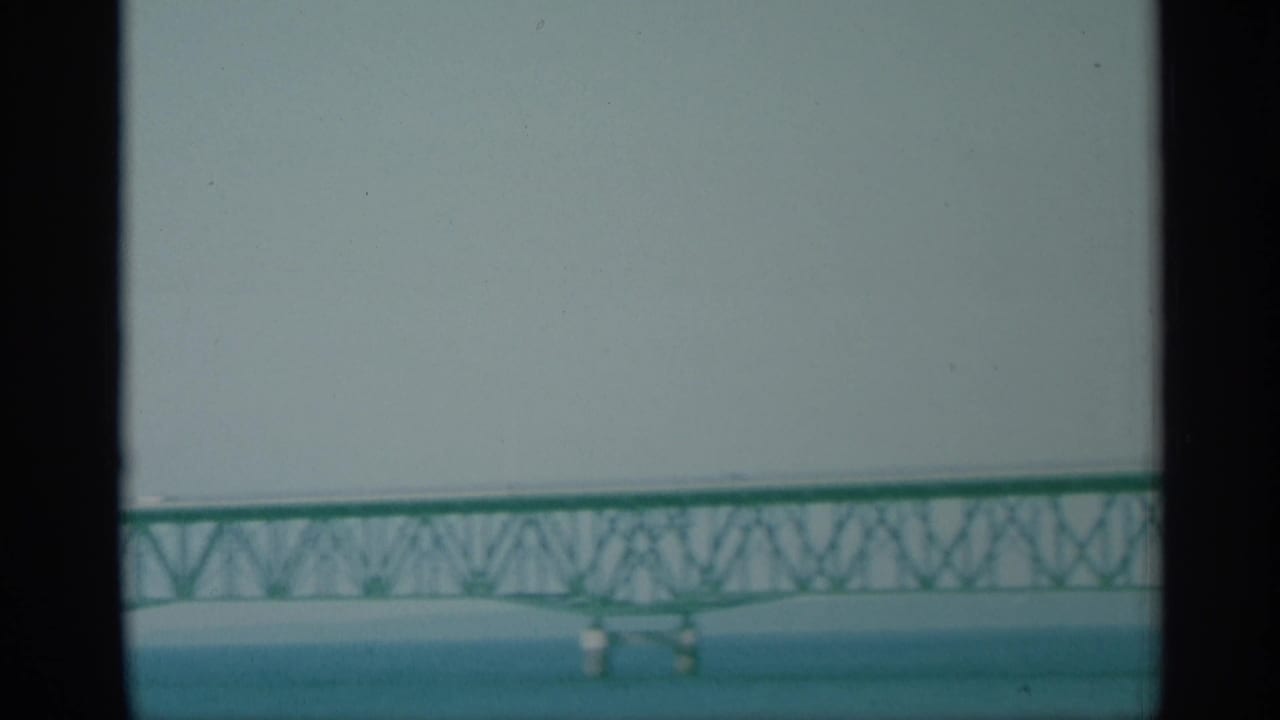 Distant 1959 New York bridge view with green truss spans, water below, and soft sky above.