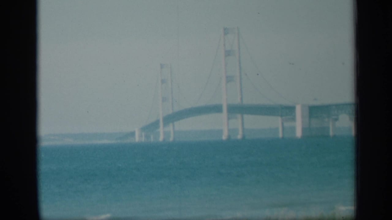 1959 New York 8mm frame showing bridge towers, cable lines, and a rising deck viewed from across the water.