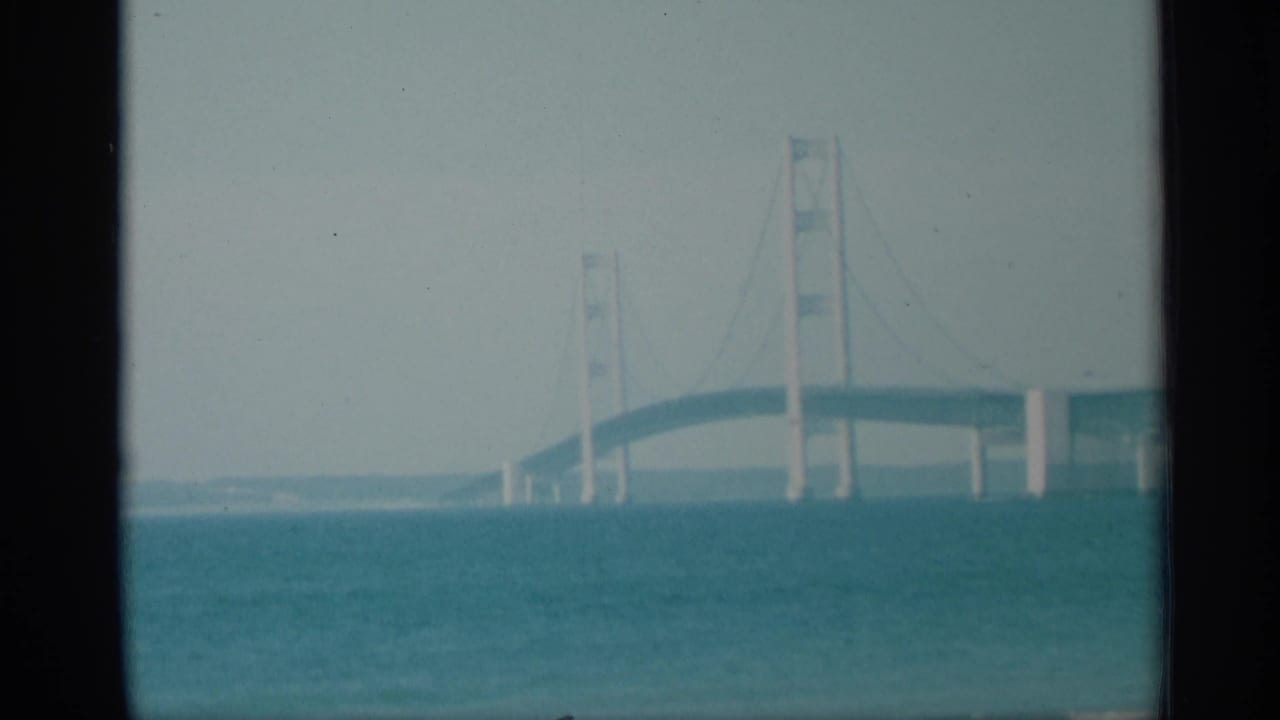 1959 New York 8mm frame showing a suspension bridge extending across blue-green water under a bright pale sky.