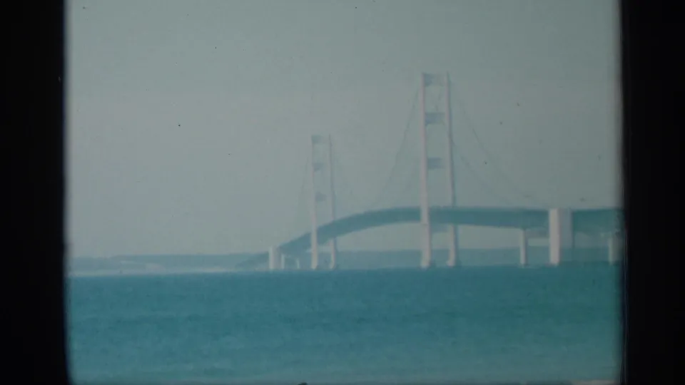 1959 New York 8mm frame showing a suspension bridge extending across blue-green water under a bright pale sky.