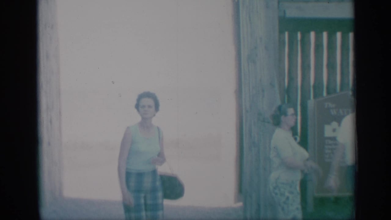 1959 8mm frame of two adult women in summer clothing near a wooden gate and posted sign at a waterfront visitor area.