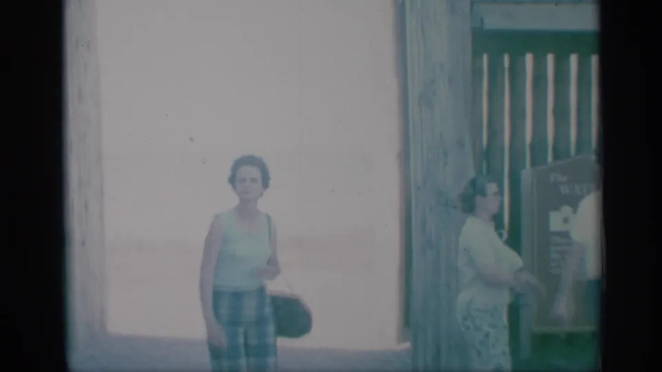 1959 8mm frame of two adult women in summer clothing near a wooden gate and posted sign at a waterfront visitor area.