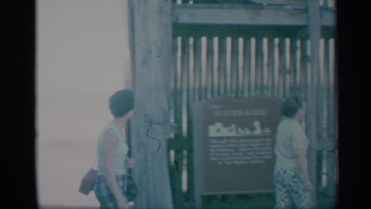 1959 8mm frame showing two adult women moving past a wooden fence and posted sign, filmed in bright washed-out light.