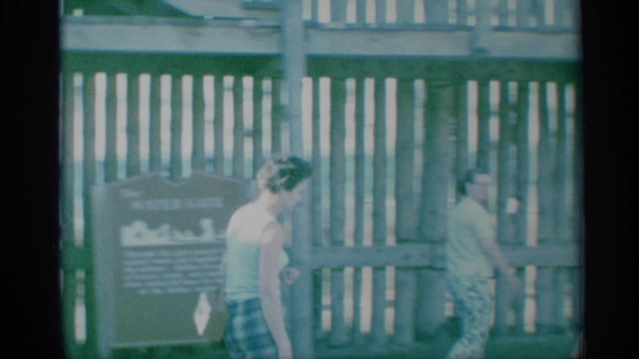 1959 8mm frame of women walking through a wooden gate beside a waterfront sign, with faded color and visible film borders.