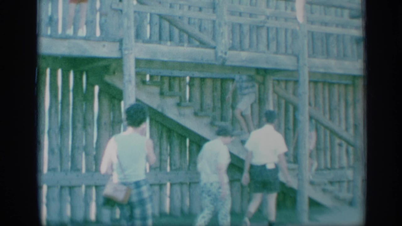 Adults in summer clothing move alongside a wooden stair and railing structure in 1959 New York home-movie footage.