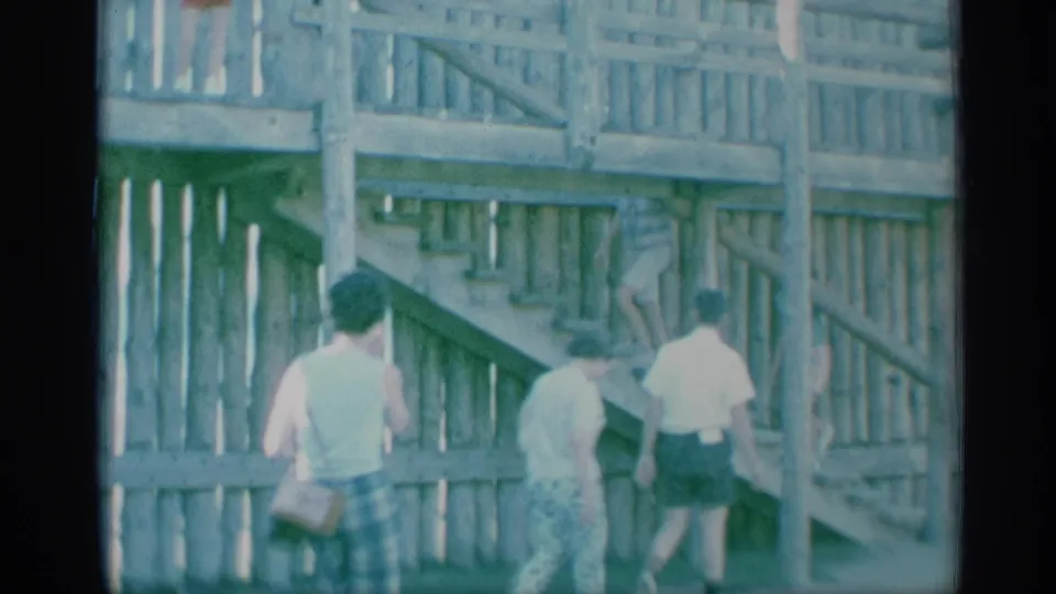 Adults in summer clothing move alongside a wooden stair and railing structure in 1959 New York home-movie footage.