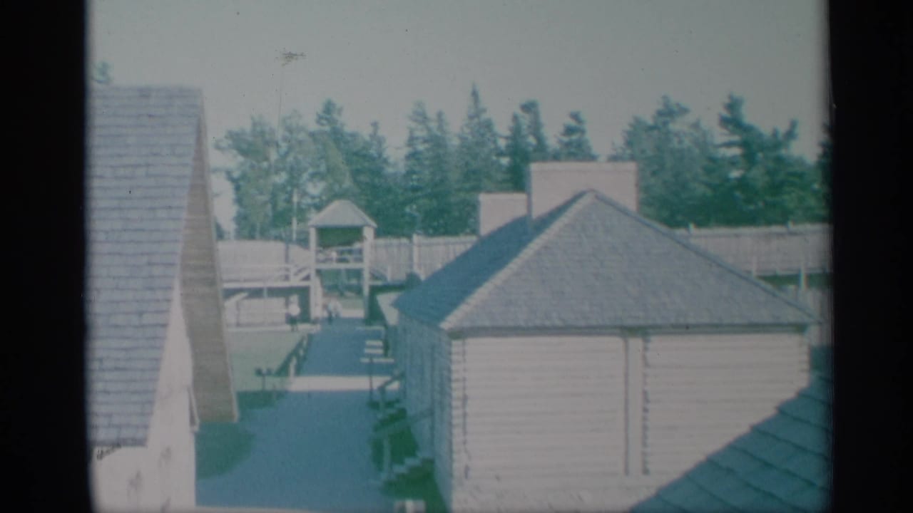 1959 New York 8mm frame showing shingled residential rooftops, a fenced yard, trees, and a small lookout-like structure near the water.