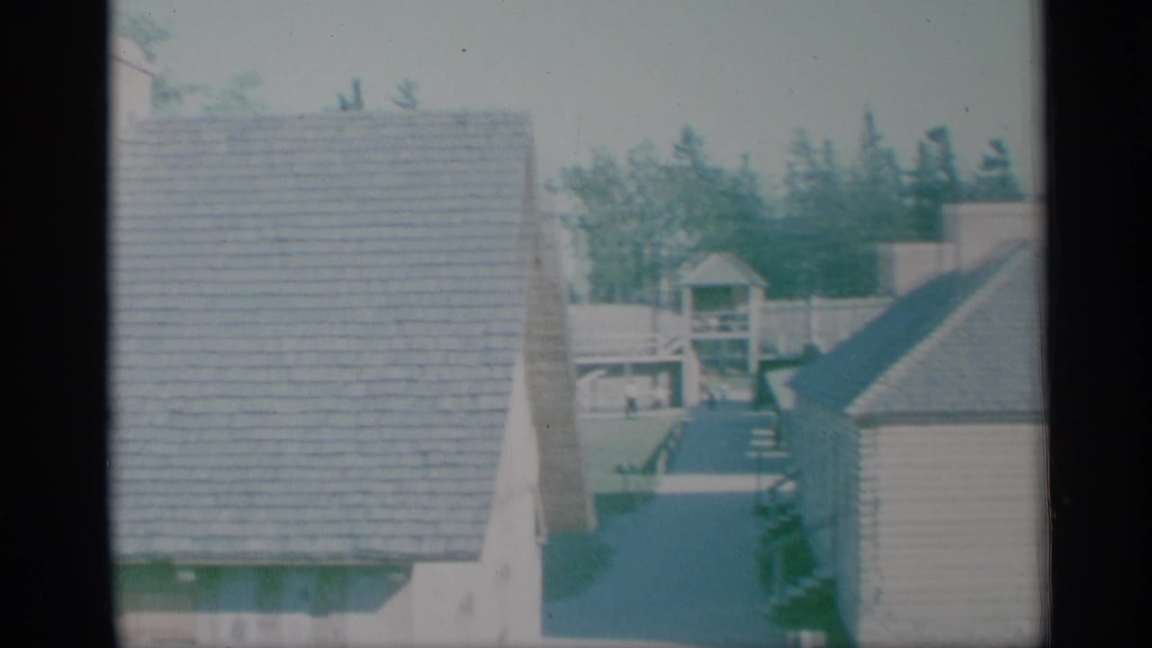 1959 New York 8mm frame with shingled houses, a fenced yard, trees, and dark foreground divisions from a visitor-shot viewpoint.
