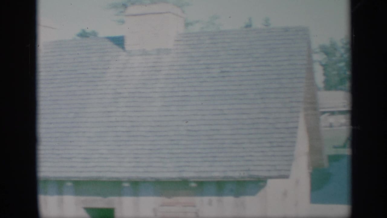 1959 New York home-movie frame showing a pale suburban house exterior with a shingled roof and partial window view.
