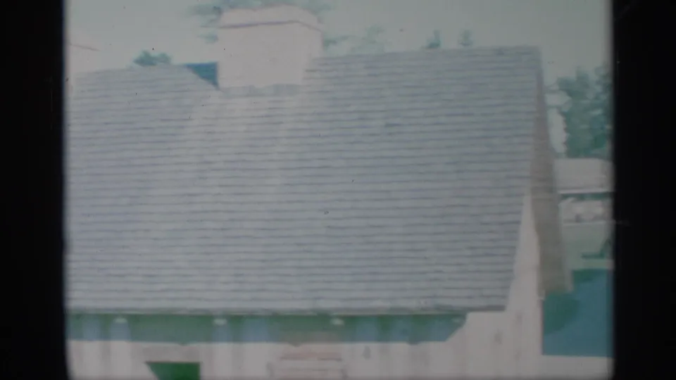 1959 New York home-movie frame showing a pale suburban house exterior with a shingled roof and partial window view.