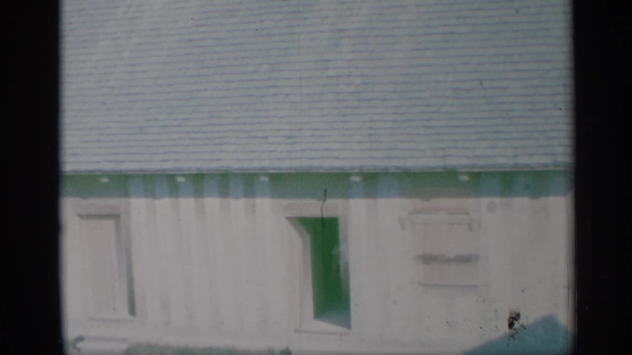 1959 New York frame of a light-colored house wall with green trim below a shingled roof and a small window.