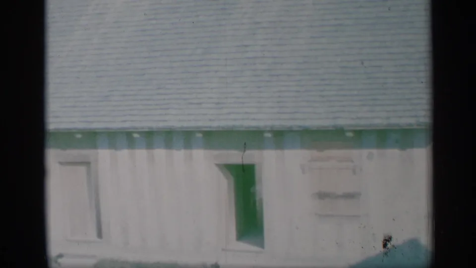 1959 New York frame of a light-colored house wall with green trim below a shingled roof and a small window.